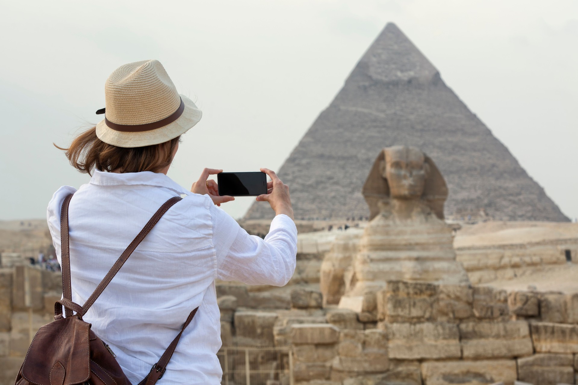 young woman standing taking a photo in front of the Great Sphinx of Giza with the amazing Pyramids in the background. Valley of the Kings, Cairo, Egypt